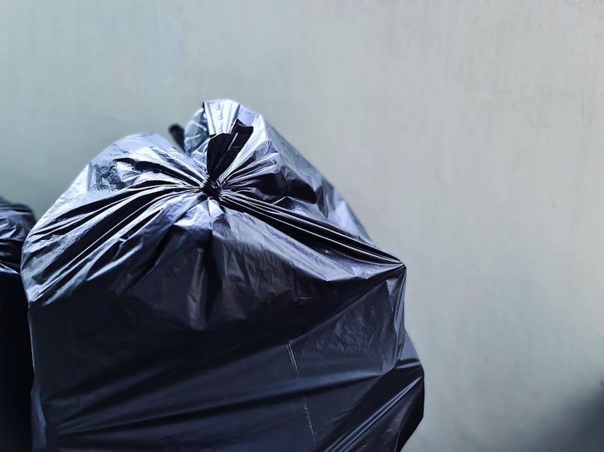 Two large black plastic garbage bags filled with waste are positioned on a concrete sidewalk, leaning against a black metal fence that runs along the edge of a residential area. The bags are crumpled 