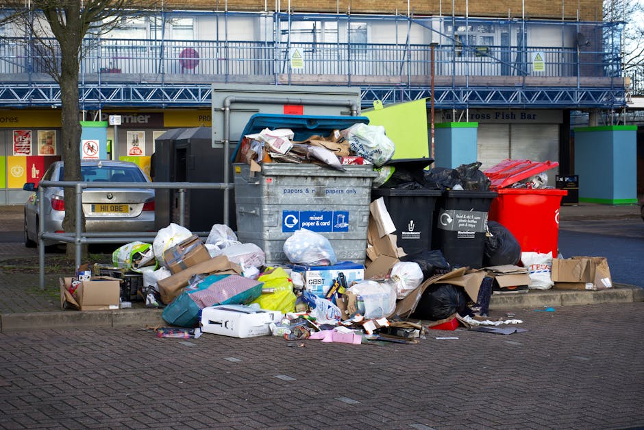 A collection of overflowing waste bins and scattered rubbish on a paved urban street, positioned on a raised curb in front of a row of shops with closed shutters. The waste includes large black garbag